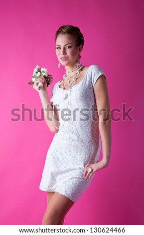 Pretty funny smiling bride girl wearing white lace wedding dress, pearls and beads, holding bunch of flowers, having fun in studio, looking at camera with happy look against pink background