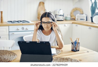 Pretty girl 12s wear headphones and glasses make task, studying online, experiences vision problems, squinting, having eyesight falls, health problems sit at table in kitchen. Poor vision, modern tech