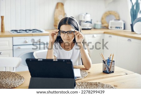Pretty girl 12s wear headphones and glasses make task, studying online, experiences vision problems, squinting, having eyesight falls, health problems sit at table in kitchen. Poor vision, modern tech