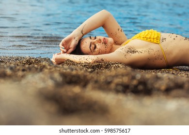 Pretty girl or beautiful woman, sexy brunette, dirty with natural sand in yellow swimsuit sleeping on sea beach with transparent water outdoors on sunny summer day on grey sandy background - Shutterstock ID 598575617