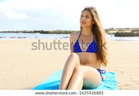 Pretty girl in blue bikini on a tropical beach. Attractive beautiful young woman sitting on beach towel on Caleton Blanco beach, Lanzarote, Canary Islands.