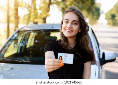 Pretty girl with cheerful smile standing near the car and showing driving license to the camera. Woman expresses her happiness after passing the driving exam. - Shutterstock ID 1921945628