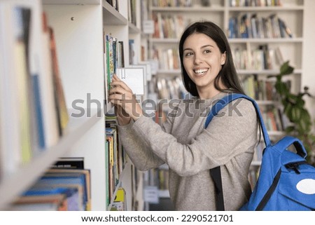 Pretty girl chooses books, standing in public library. Caucasian female student prepares for university admission looks interested and motivated, take textbook staring aside. Education, new knowledge