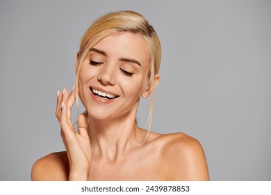 pretty girl with clear skin smiling broadly and touching skin with pleasure against gray background - Powered by Shutterstock