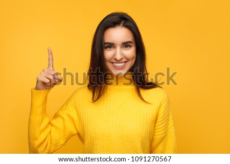 Pretty girl in colorful sweater holding forefinger up smiling at camera on yellow background. 