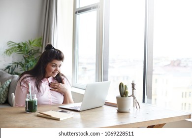 Pretty girl with curvy body and dark hair smiling excitedly, holding hand on her chest and looking at laptop screen, reading astonishing news online, surfing internet, sitting at window. Copy space
