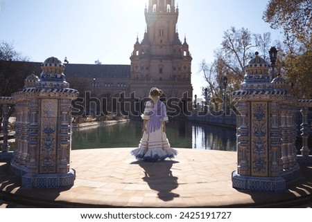 A pretty girl dancing flamenco in a frilly and fringed dress in a famous square in seville, spain. The girl is wearing a flower in her hair and her back is turned. In the background a tower