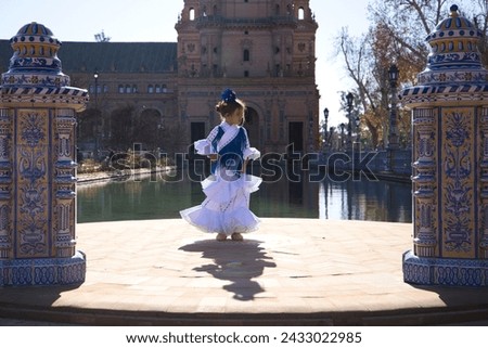 A pretty girl dancing flamenco in a frilly and fringed dress in a famous square in seville, spain. The girl is wearing a flower in her hair. In the background a tower