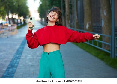 Pretty Girl Dancing On The Street With Phone, Cute Millenial Woman In Red Stylish Sweater Having Smartphone Dance At Modern City Background