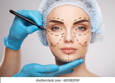 Pretty girl with dark eyebrows wearing blue medical hat at studio background, doctor's hands wearing blue gloves drawing perforation lines on face, plastic surgery concept. - Shutterstock ID 605382959