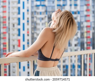 Pretty girl with freckles in a purple lace top and sweatpants. Stands on the balcony with his head thrown back. Against the background of a multi-colored multi-storey building.