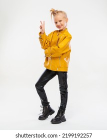 Pretty Girl Gesture V Sign For Victory Or Peace Sign In A Leather Mustard Jacket, Black Snake Texture Leggings And Boots. High Fashion, Full Length Standing Pose, Isolated Against A Studio Background