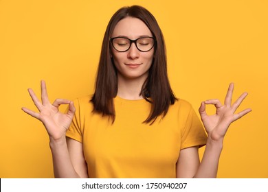Pretty Girl In Glasses Feel Relaxed, Joyful And Calm. Attractive Peaceful Young Woman With Closed Eyes Gather Patience, Breathing Air, Holding Hands In Zen Mudra Gesture. Meditation, Practice Yoga