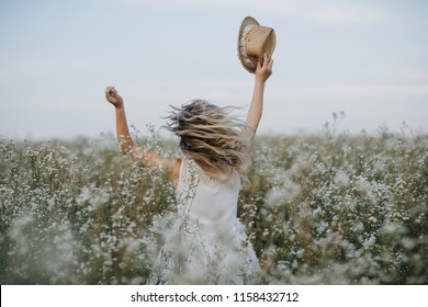 Pretty girl with a hat in her hand walks in a field with field flowers and smiles sincerely - Powered by Shutterstock