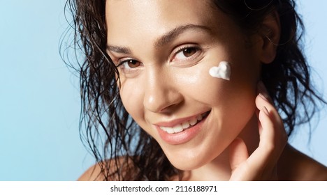 Pretty  girl with heart shaped face cream or foam on cheek, healthy skin, smiling during daily skincare routine, looking at camera on blue studio background. Natural woman beauty concept.