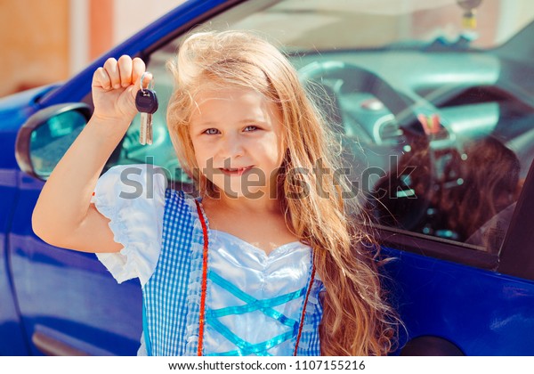 Pretty girl with\
long golden hair wearing princess dress and showing keys standing\
near new blue car in\
sunlight