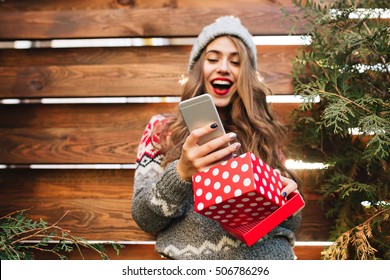 Pretty Girl With Long Hair With Christmas Box On Wooden Background. She Wears Warm Winter Clothes, Looks Astonished At Phone In Hand