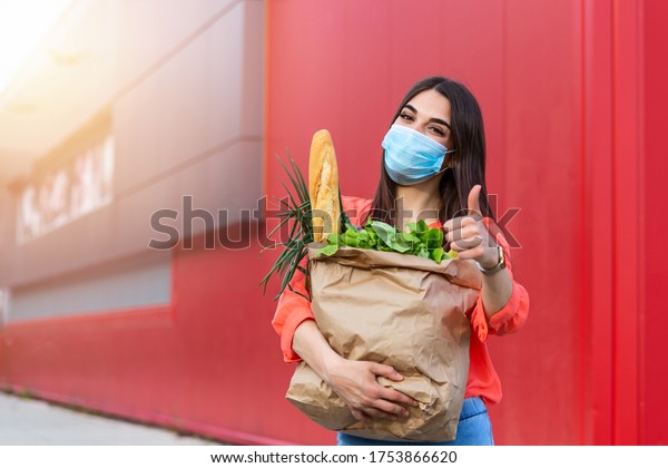 Pretty girl with medical mask holding bag with\
groceries and looking at camera with thumb up. Woman holding heavy\
bag with groceries. Groceries shopping during Covid 19, coronavirus\
pandemic