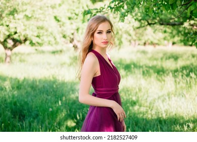 Pretty Girl With Professional Makeup And Curls In A Violet Purple Dress Looks Directly Into The Camera And Poses. Bridesmaid In The Garden. High Quality Shot