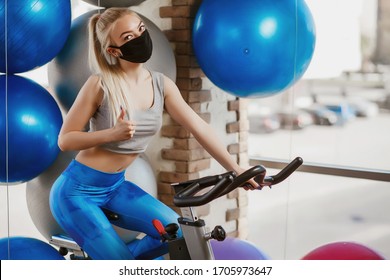 A pretty girl in a protective mask is engaged in an exercise bike. Protective masks against virus infection. Training during quarantine in the gym. Coronavirus (COVID-19) protection concept. - Powered by Shutterstock