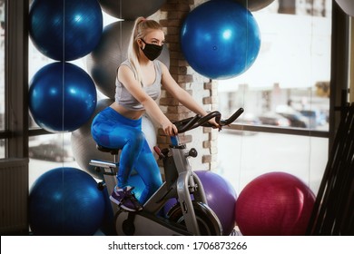 A pretty girl in a protective mask is engaged in an exercise bike. Protective masks against virus infection. Training during quarantine in the gym. Coronavirus (COVID-19) protection concept. - Powered by Shutterstock