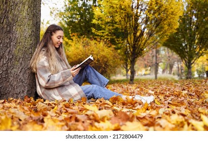 Pretty girl reads book in autumn park. Young woman sits on background of nature. Rest pastime leisure