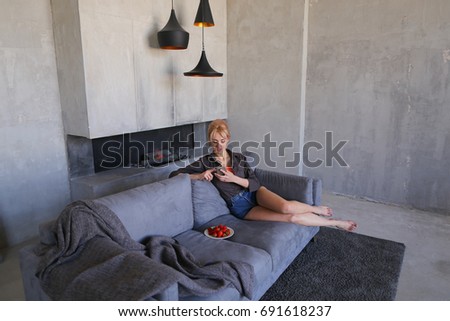 Pretty girl rests from bustle of city and sits on Internet with mobile phone, looks ahead and tries ripe red strawberry berries, sits on gray soft sofa in stylish living room with gray walls. Young