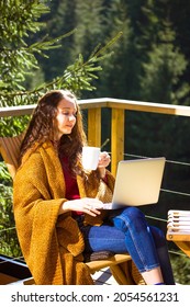 Pretty girl sitting on balcony terrace wrapped in yellow plaid holding cup of tea coffee, working remotely at laptop. Young brunette woman shopping online. House in pine forest at sunny autumn day.