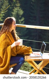 Pretty girl sitting on balcony terrace wrapped in yellow plaid holding cup of tea coffee, working on laptop. Brunette woman works remotely, shopping online. Woodland house in pine wood at autumn day.