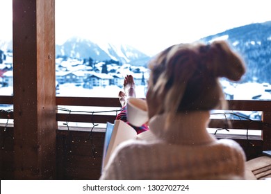 Pretty Girl Sitting On The Wooden Balcony In A Hotel, Put Her Long Legs On The Handrail And Drinking Morning Latte. Wearing Pajama And Sweater. Enjoys Amazing Snowy Mountains View. Chilling Out