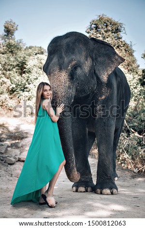 Pretty girl in sunglasses and green dress print kindly strokes elephant’s trunk; exotic animals, glamour.