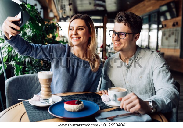 カフェでコーヒーを飲みながら ボーイフレンドと一緒にスマートフォンを使って自撮りをする美しい女の子 の写真素材 今すぐ編集