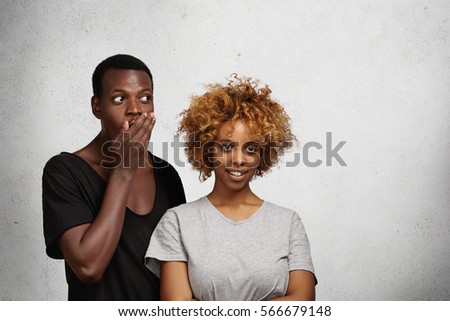 Pretty girl with trendy hairstyle posing indoors with happy smile while African man wearing blackt-shirt with low neck standing beside her, covering mouth with his hand and looking away with shock