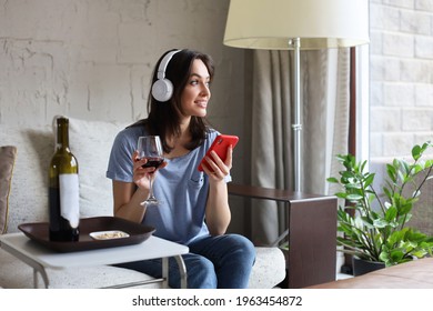 Pretty girl using her smartphone on couch at home in the living room. Listening music, drinking red wine, relaxation after a hard week at work - Shutterstock ID 1963454872