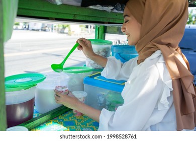 a pretty girl in a veil sells es campur using a scoop to get coconut milk from a jar on a cart