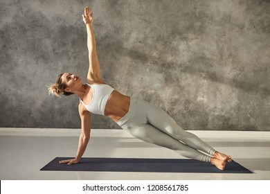 Pretty girl wearing leggings and short top standing in side plank on one hand at gym, training body core and balance, strengthening abs muscles. Attractive female doing planking bodyweight exercise