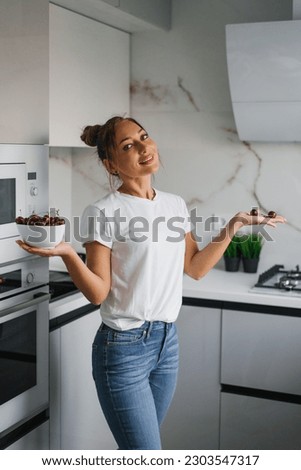 Pretty girl in a white T-shirt is standing in the kitchen and holding a white vase with red, ripe cherries. The concept of healthy food