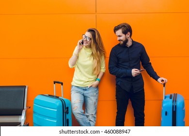 Pretty girl in yellow sweater with long hair is speaking on phone on orange background. Handsome guy with beard is looking at her near. They have two suitcases.