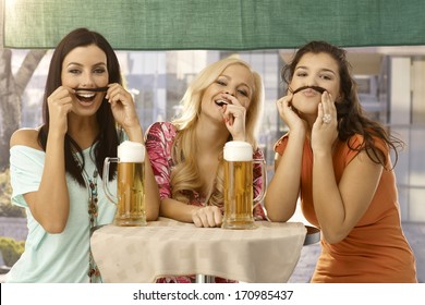 Pretty girls having fun, forming moustache from hair, drinking beer in outdoor bar, smiling.