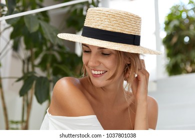 Pretty glad female wears straw hat, shows her tanned bare shoulder, smiles with happy expression as looks joyfully aside, spends summer vacations abroad. Lovely young woman enjoys good resort alone