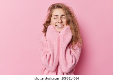 Pretty Glad Woman With Long Wavy Hair Closes Eyes From Enjoyment Wears New Cashmere Jumper Keeps Hands Under Chin Smiles Gently Isolated Over Pink Background. Happy Emotions And Coziness Concept