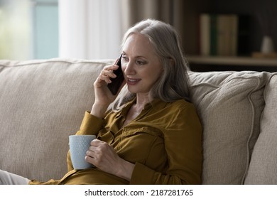 Pretty Grey Haired Retired Grandma Woman Talking On Mobile Phone, Making Cellphone Call To Family, Grandchildren, Speaking, Listening, Smiling, Drinking Cup Of Hot Tea, Coffee