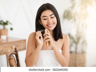 Pretty Happy Asian Woman Applying Hair Oil To Prevent Split Ends, Hair Care At Home Concept, Copy Space. Beautiful Lady Wrapped In Towel Using Hair Spray, Hair Treatment At Home Concept