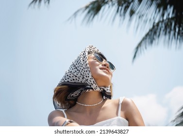 Pretty happy Asian woman portrait in casual white shirt wearing sunglasses and hair scarf, enjoy with sunshine at the beach under the coconut palm trees and blue sky background in summer.