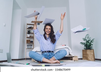 Pretty Happy Excited Female Student Sitting On Floor With Crossed Legs And Raised Hands Throwing Up Papers After Finishing Hard Task