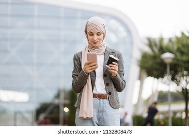 Pretty happy Muslim lady in hijab smiling at camera, standing outdoors with coffee to go and phone in hands. Likable Arab woman in headscarf walking on the street with take away coffee and phone - Powered by Shutterstock