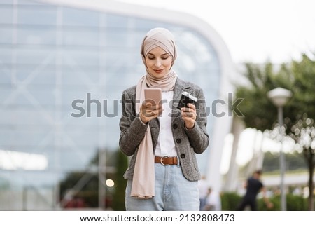 Pretty happy Muslim lady in hijab smiling at camera, standing outdoors with coffee to go and phone in hands. Likable Arab woman in headscarf walking on the street with take away coffee and phone