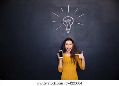 Pretty happy young woman holding and pointing on smartphone blank screen standing over chalkboard background - Powered by Shutterstock