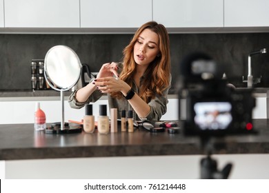 Pretty healthy young girl recording her video blog episode about new cosmetic products while sitting at the kitchen table at home and applying make-up