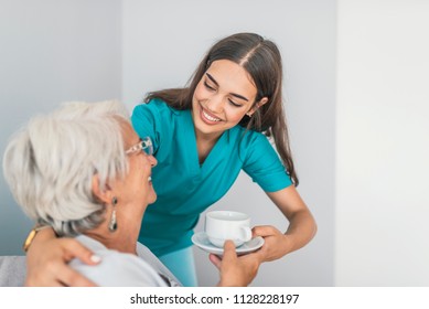 Pretty Helpful Carer Talking With Female Patient. Health Visitor And A Senior Woman During Home Visit. Supportive Senior Care Assistant And Smiling Older Woman In Nursing Home